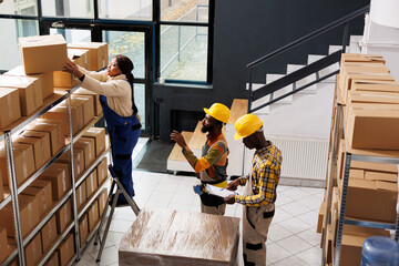 African american post office employee standing on ladder and taking parcel from shelf. All black package handlers team inspecting order and preparing cardboard box for shipment top view © DC Studio