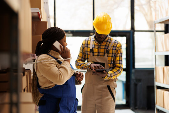 Warehouse worker checking parcel in tablet and manager answering supervisor call. African american woman talking on landline phone and assistant using inventory software in storehouse