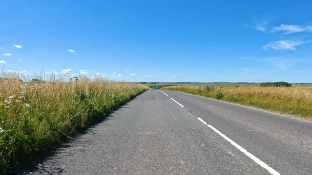 Traveling by car on an asphalt road through the beautiful English countryside. Left hand drive past green fields and pastures spread across the picturesque rolling landscape on a sunny summer day.
