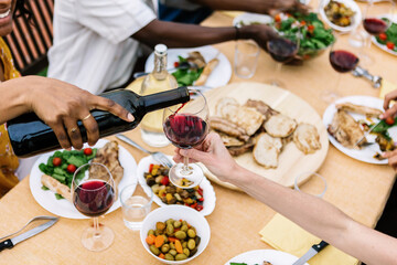 Young woman pouring a glass of wine to her friend at dinner summer party. Millennial people enjoying time together sitting on table at rooftop celebration.