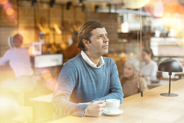 Pensive middle-aged man in casual clothes with mug of hot drinks, enjoying tea or coffee at cafe...