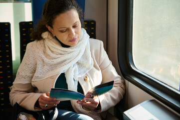 Confident mid adult multi ethnic woman enjoying train ride and reading a leaflet or book, sitting...