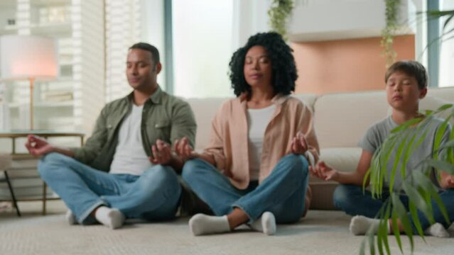 Calm family multiracial African American mother and father meditate with kid boy child yoga exercise in lotus pose on home floor together parents teaching little son zen balance breathing meditation