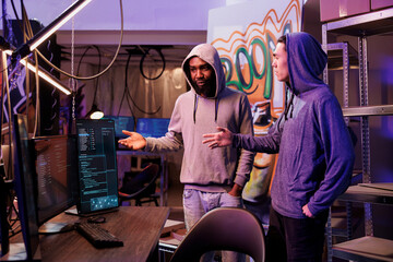 Hackers discussing hacking plan and cybercrime technique. Criminals in hood standing near computer and talking about malicious software code on screen in abandoned warehouse