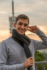 Young brown haired man smiling at the camera while touching his ear during sunset. Vertical