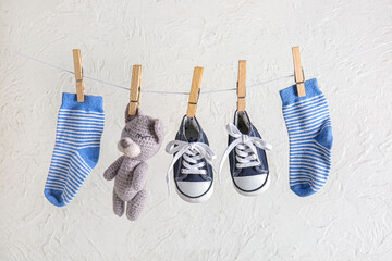 Stylish baby shoes with cute toy and socks hanging on rope against grunge white background