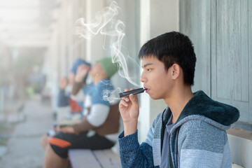 Asian preteen sitting in front of windows of school building and smoking e-cigarette not far from his friends, bad habit and behavior of young people in drug concept, soft focus, white smoke edited.