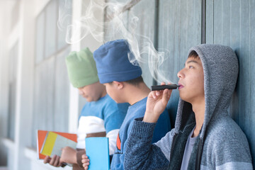 Asian preteen sitting in front of windows of school building and smoking e-cigarette not far from his friends, bad habit and behavior of young people in drug concept, soft focus, white smoke edited. - obrazy, fototapety, plakaty