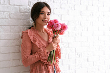 Beautiful young Asian woman with bouquet of pink dahlias near white brick wall
