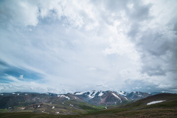 Dramatic alpine view from stony grassy hill to large snow-capped mountain range under cloudy gray sky. Beautiful mountain landscape with green rock hills against high snowy mountains in rainy weather.