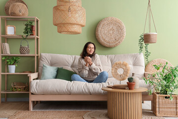 Pretty young woman with cup of tea resting on cozy white sofa in living room