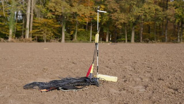 High Resolution Video of a Geophone standing on a field. Next to the Geophone are cables, going into the earth to measure the reflections of the seismic vibration. You can see trees in the background.