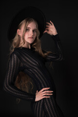 Stunning portrait features slim blonde fashion model exuding sensuality in relaxed pose on black background. Woman holds brim of hat with one hand and puts other hand on waist, dressed in black dress