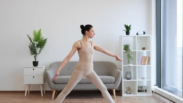 Mature brunette practising Warrior One Pose during yoga exercise at home. Caucasian woman spread her legs wide and pulls one knee forward, straightening arms up above the head.