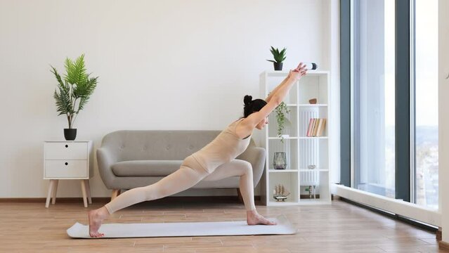 Mature brunette practising Warrior I Halfway Fold during yoga exercise at home. Caucasian woman spread her legs wide and pulls one knee forward, straightening arms up above the head.