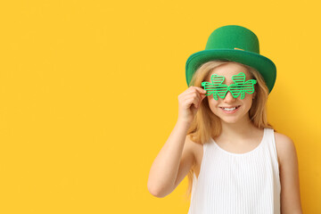 Cute little girl in leprechaun hat and decorative glasses in shape of clover on yellow background. St. Patrick's Day celebration