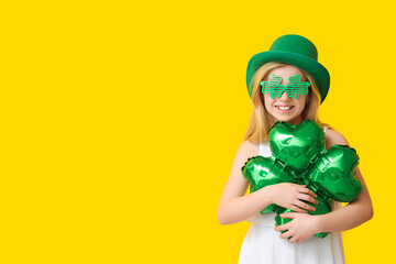 Cute little girl in leprechaun hat and decorative glasses with air balloon in shape of clover on yellow background. St. Patrick's Day celebration