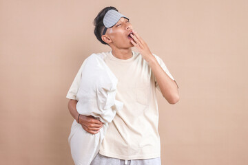 Sleepy young Asian man with pajama and eye mask yawning while holding a pillow  