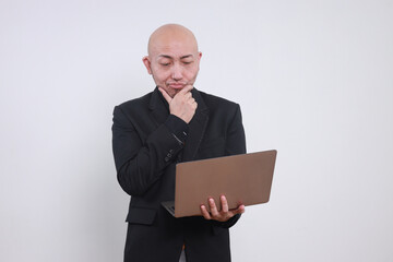 Adult Asian businessman holds chin, purses lips and looking at laptop computer thinking.