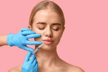 Young woman receiving filler injection in lips on pink background
