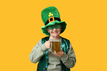 Senior woman in leprechaun hat and decorative glasses with glass of beer on yellow background. St....