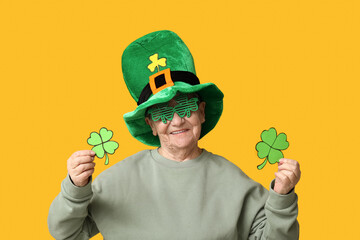 Senior woman in leprechaun hat and decorative glasses with clover on yellow background. St....