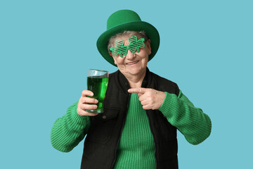 Senior woman in leprechaun hat and decorative glasses pointing at glass of beer on blue background....