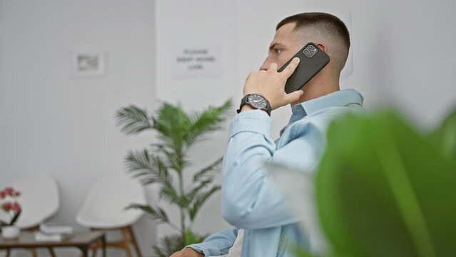 Hispanic man talking on smartphone in lobby with watch, indoor foliage, and chairs.