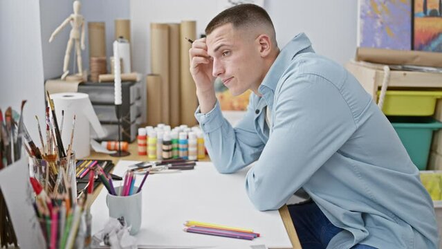 A contemplative man in a blue shirt sits in a creative studio with art supplies, reflecting on his work.