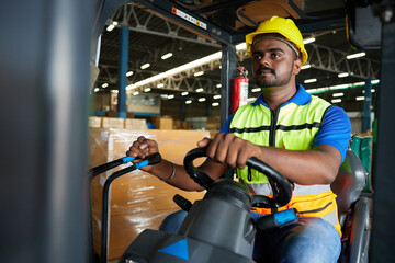 Indian worker driving a forklift and looking to something in warehouse storage