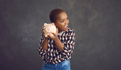 Studio portrait of happy smiling black woman holding pink piggy bank with savings to her ear, listening to sound of coins clinking inside, anticipating spending whole lot of money on fulfilling dreams - obrazy, fototapety, plakaty