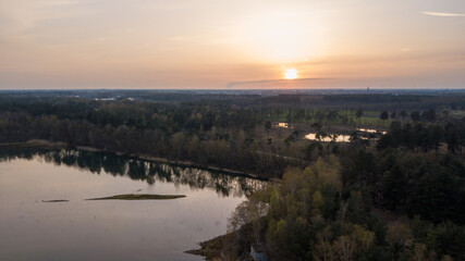 An aerial view captures the serene beauty of a forest lake at twilight. The setting sun dips towards the horizon, casting a gentle golden glow across the water's surface and the surrounding trees. The