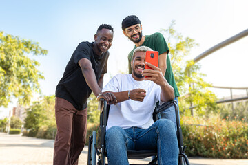 Fototapeta premium Young African American man in a wheelchair using phone together with male friends outdoors.