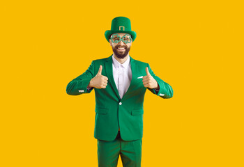 Happy bearded man enjoying cool great party on St Patrick's Day. Cheerful joyful man in green costume, hat and funny shamrock clover glasses smiles and gives thumbs up isolated on yellow background - obrazy, fototapety, plakaty