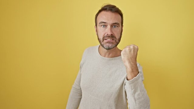 Frustrated mid-aged hispanic man, fists clenched, seething with pure fury. mad, aggressive stand against yellow wall. face shows a serious, upset expression