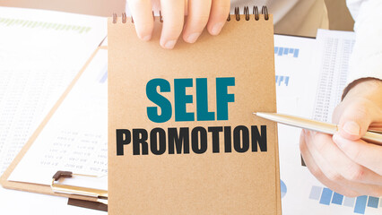 Text SELF PROMOTION on brown paper notepad in businessman hands on the table with diagram. Business...