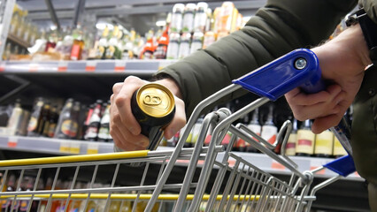Close-up of a shopping trolley in alcohol department and a male buyer's hand putting a golden-black...
