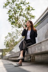 Fototapeta premium Asian businesswoman sitting and using phone outdoor, with a tumbler beside her