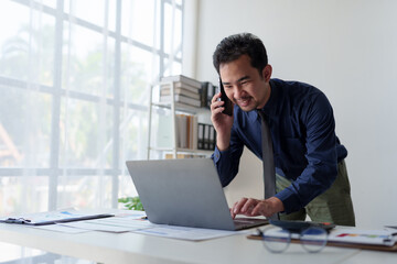 Fototapeta premium Businessman relaxes holding smartphone, checking email, work details on laptop computer online, contacting, business negotiation with customer pointing to successful business goals in office.