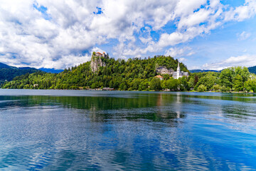 Fototapeta na wymiar Scenic view of Lake Bled with beautiful reflections and castle on a rock and woodland in the background on a blue cloudy summer day. Photo taken August 8th, 2023, Bled, Slovenia.