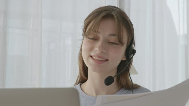 A nice young woman smiling and talking into her headset. Using computer and documents as she talks
