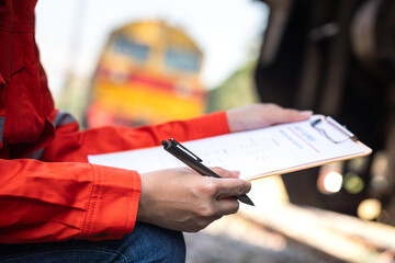 A service technician is checking on heavy machine maintenance checklist, with an ancient train locomotive head as blurred background. Transportation industrial working scene, selective focus at hand.