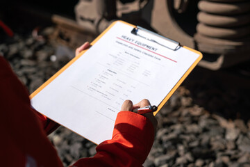 Action of a mechanic engineer is checking on heavy machine checklist form to verify the quality of maintenance service, with train locomotive part as blurred background. Industrial working scene.