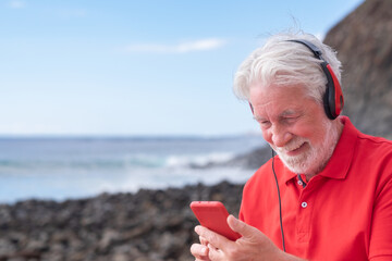 Smiling relaxed bearded senior man with red clothes in outdoors at the beach using mobile phone,...