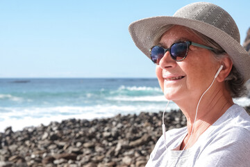 Smiling relaxed senior pensioner woman with hat sitting in outdoors at the beach listening music by earphones, elderly female enjoying free time and retirement