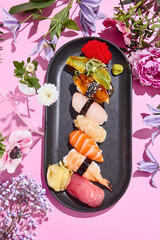 Elegant sushi selection with tuna, shrimp, salmon, sea bass, eel and avocado on a stylish black serving plate against a pink background, with surreal floral accents
