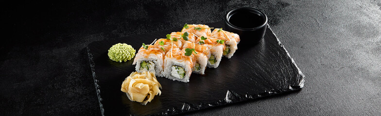 Shrimp roll with shrimp on top and cucumber and cheese inside, on a black slate with soy sauce, wasabi, and ginger