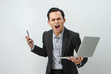 Angry mad asian indonesian business man in suit holding smart phone and laptop computer on isolated...