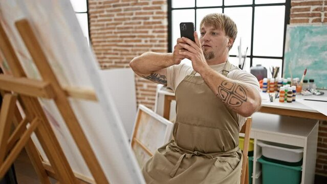 A focused man in an apron uses a smartphone in a sunny art studio with easels and paint supplies.