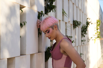 Young gay boy with pink hair and make-up leaning against a white wall with rectangular holes in...